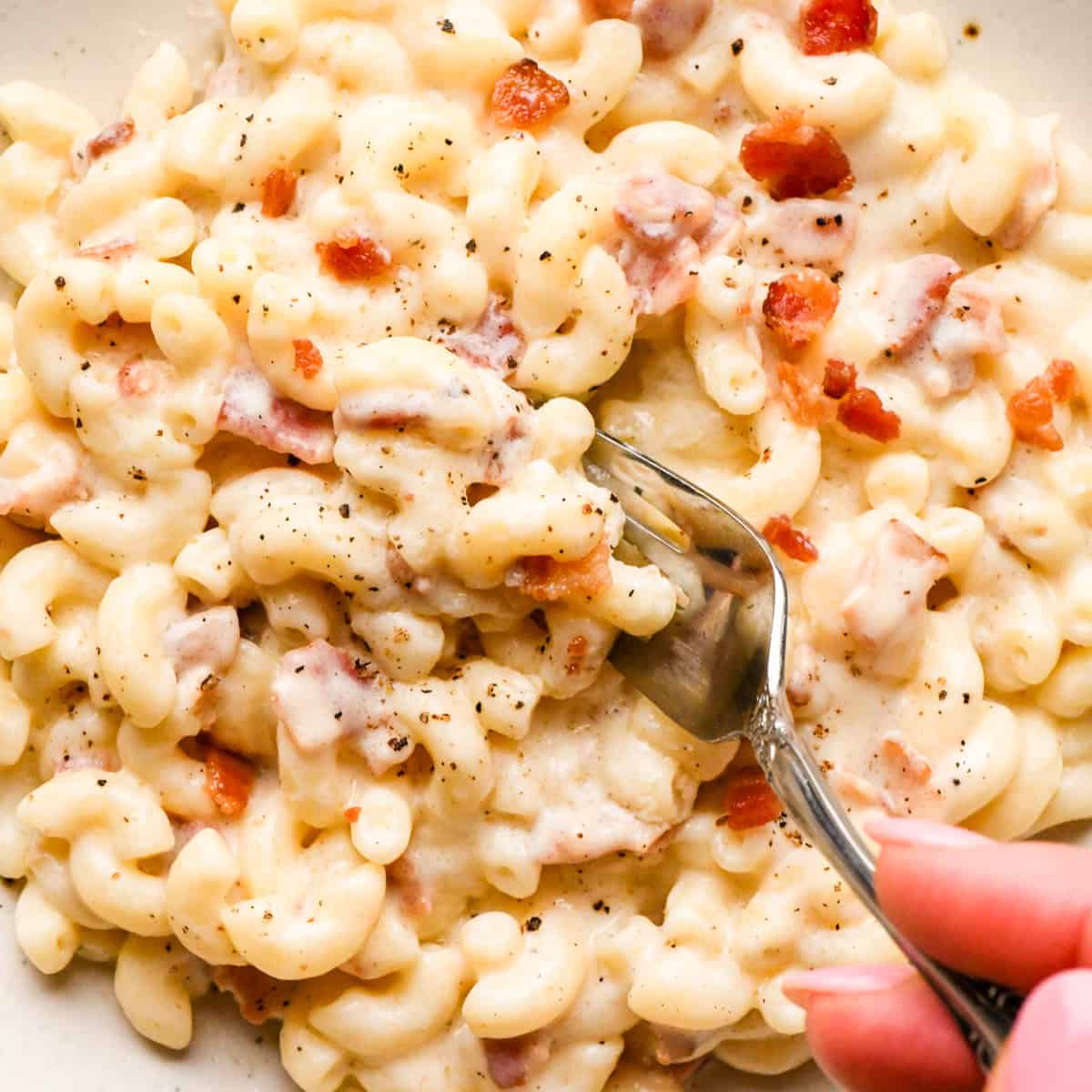 close up photo of a hand holding a fork taking a bite of  Bacon macaroni and cheese on a white speckled plate, topped with cooked bacon pieces and freshly ground black pepper