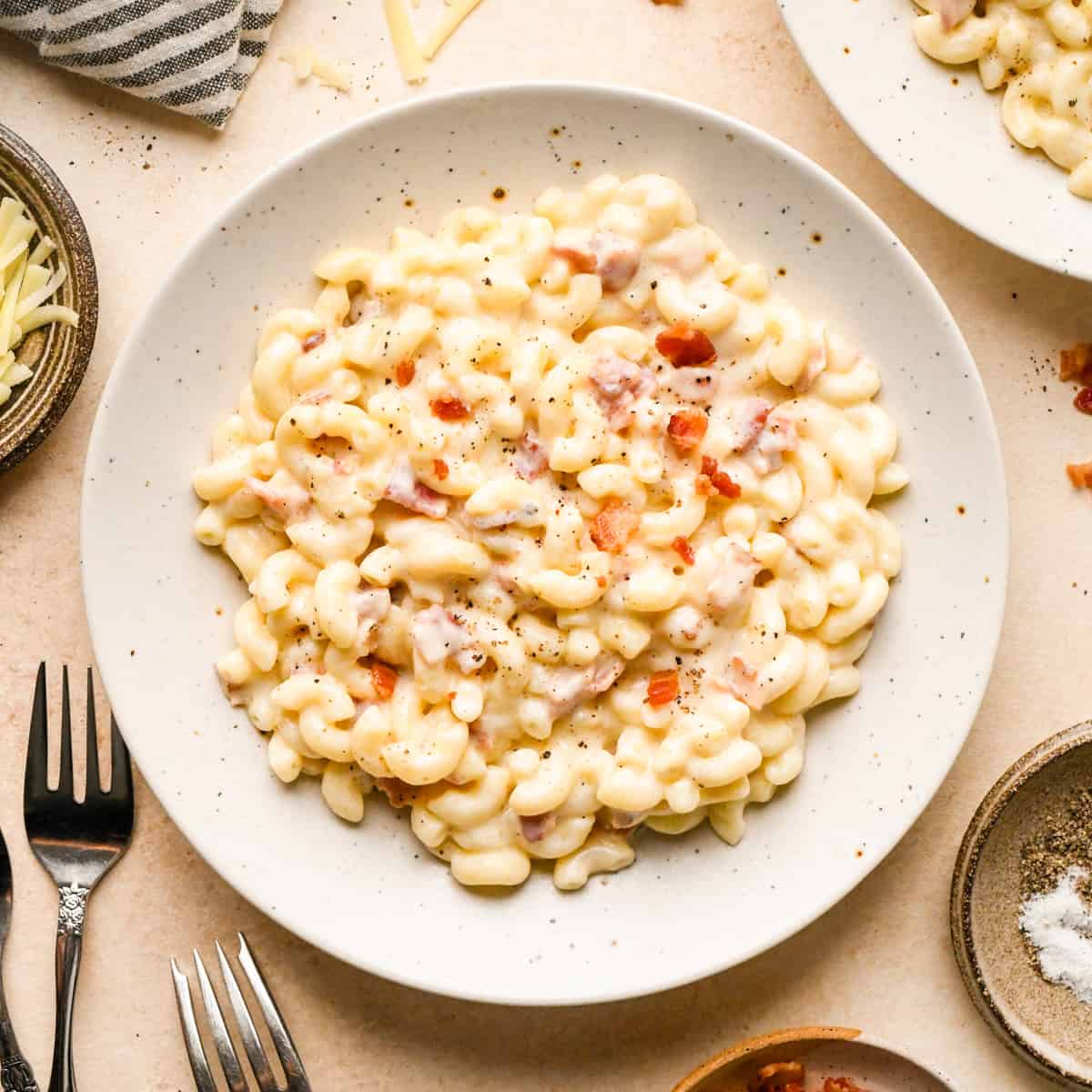 Bacon macaroni and cheese on a white speckled plate, topped with cooked bacon pieces and freshly ground black pepper, surrounded by another bowl of bacon mac and cheese, a bowl of shredded cheese, a bowl of salt and pepper and two forks