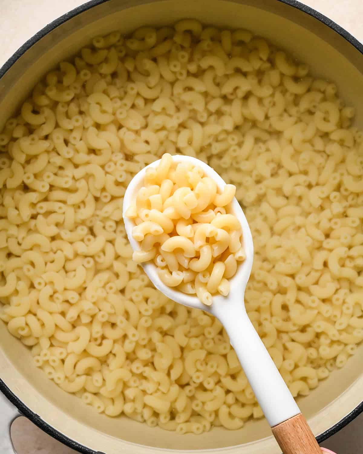a white and wood spoon holding cooked macaroni over a pot of macaroni in water to make bacon mac and cheese