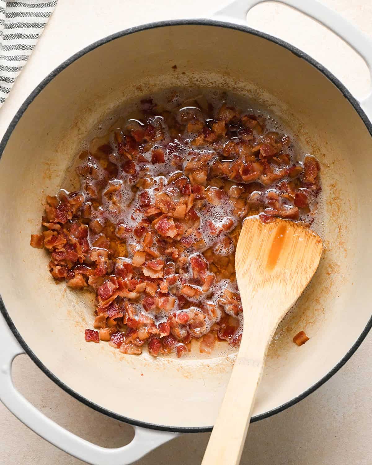 diced bacon after it has been cooked in a white pot with a black rim and a wooden spoon