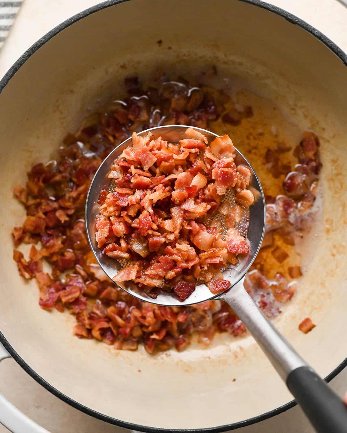 a mesh metal spoon with cooked bacon in it being held over a white pot with more cooked bacon and bacon grease making bacon macaroni and cheese