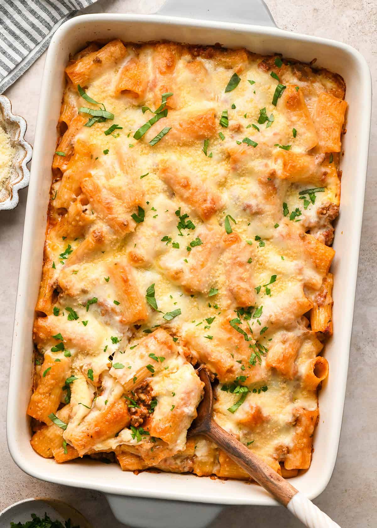 Baked ziti in a white casserole dish topped with fresh herbs, being served with a wooden spoon.