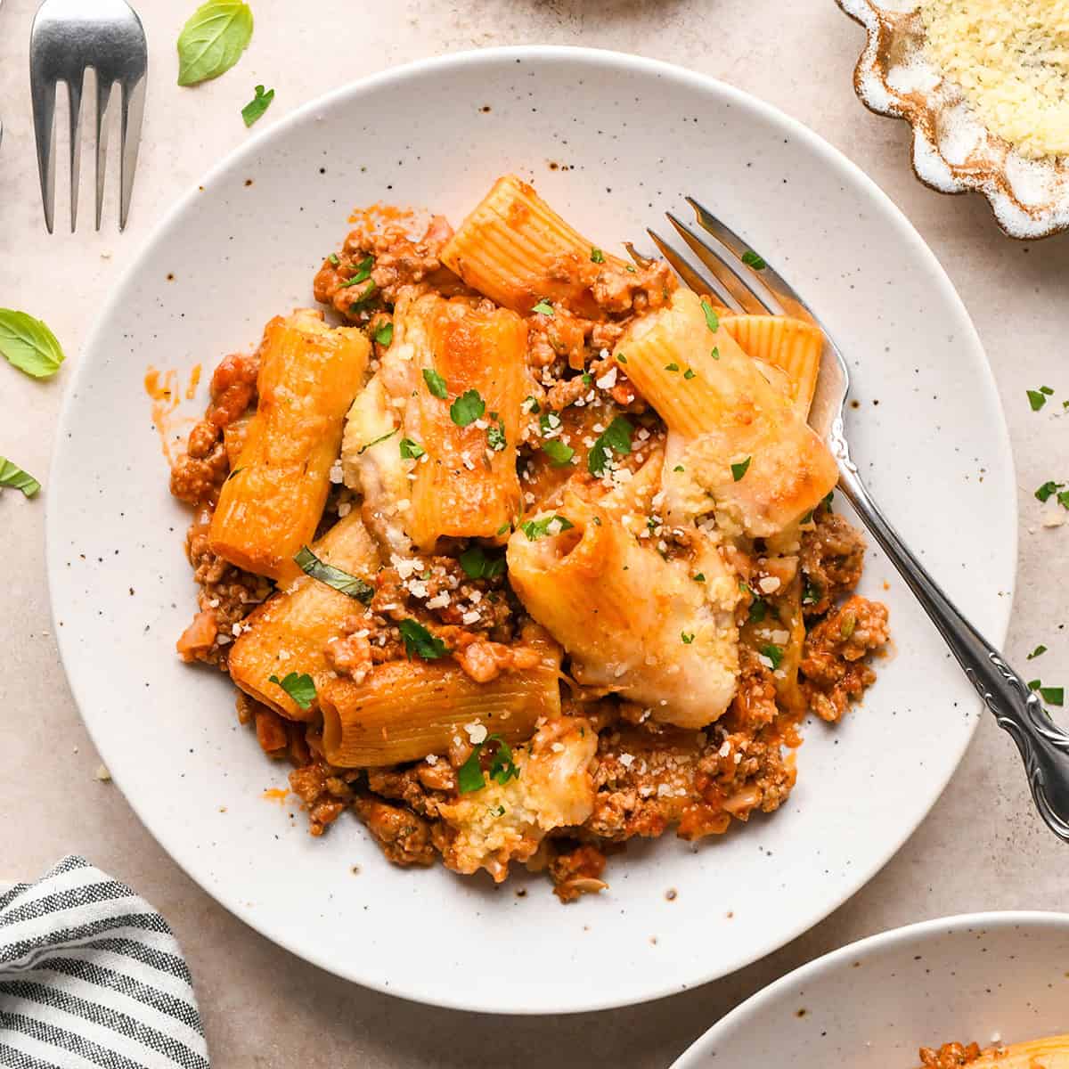 Close-up photo of baked ziti topped with fresh herbs, served on a white speckled plate with a fork.
