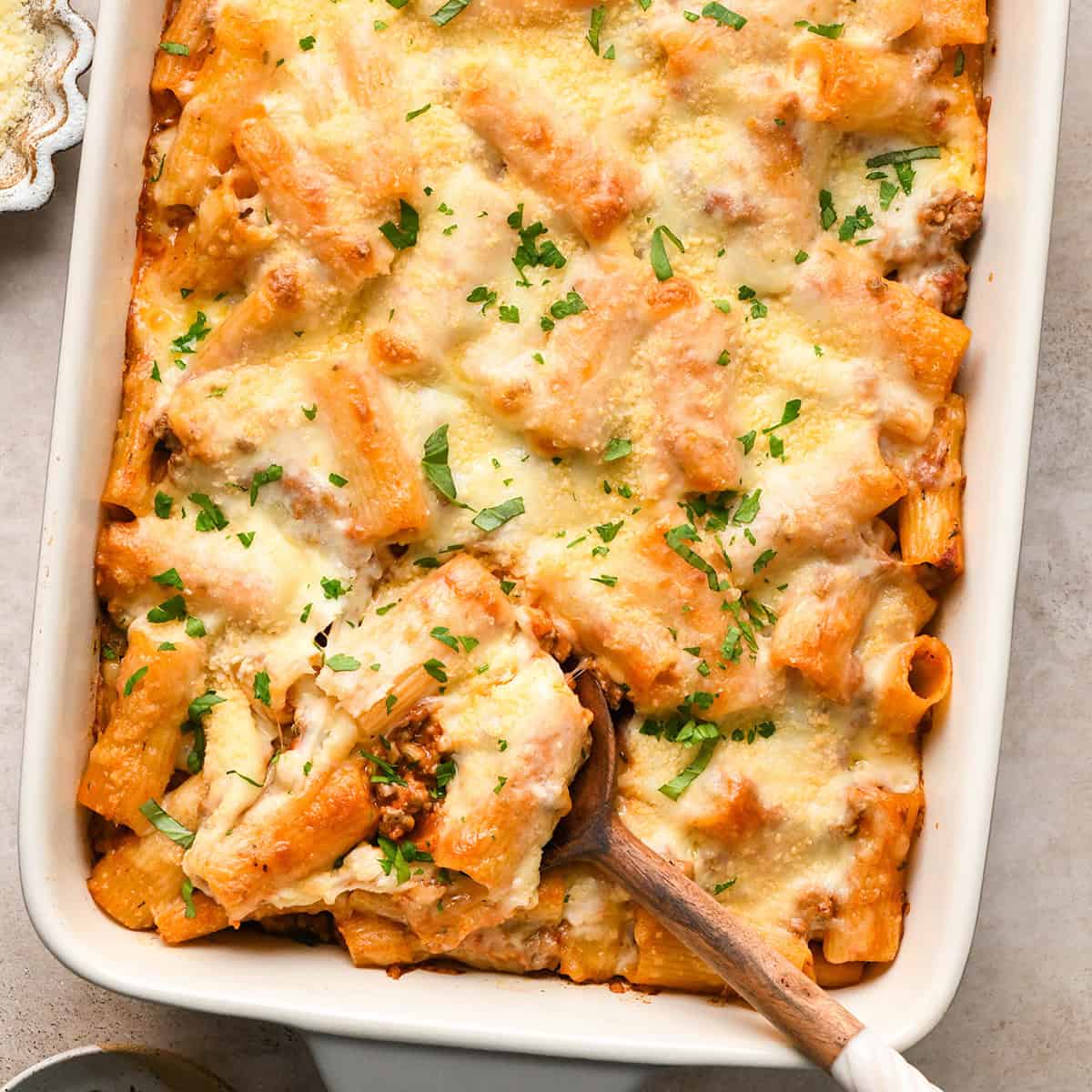 Baked Ziti in a baking dish garnished with parsley, a spoon is taking a scoop out of the dish