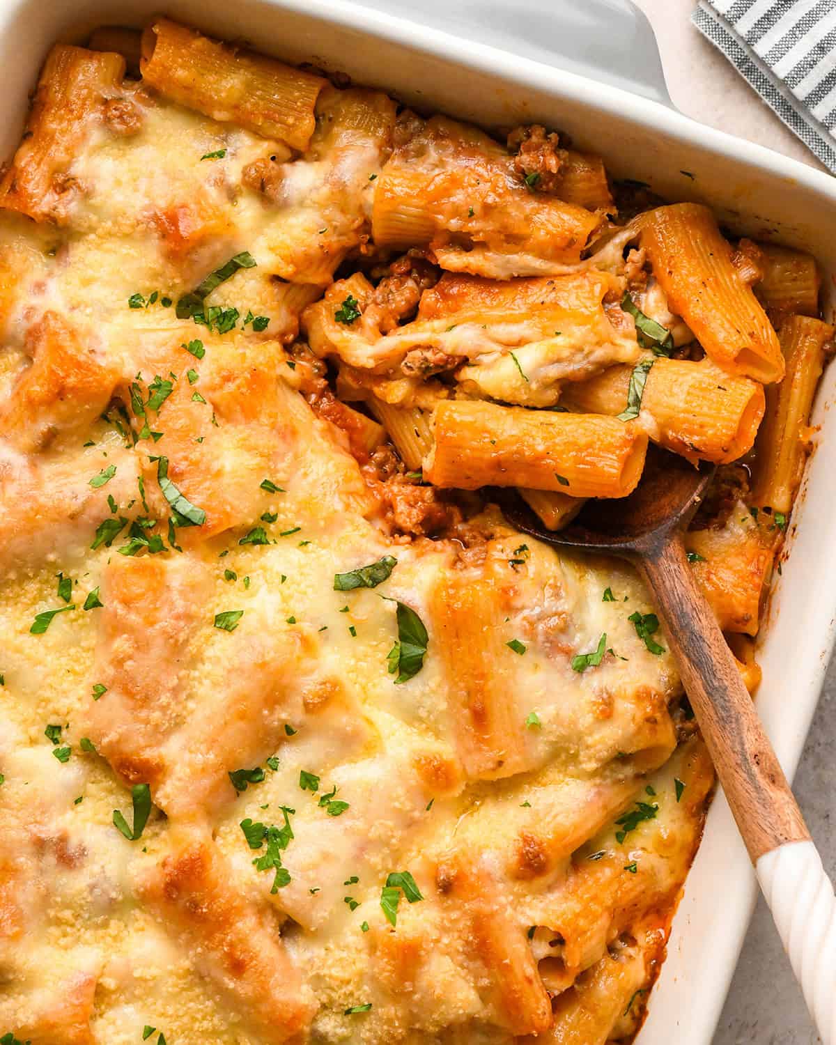 Baked Ziti in a white baking dish garnished with parsley, a spoon is taking a scoop out of the dish