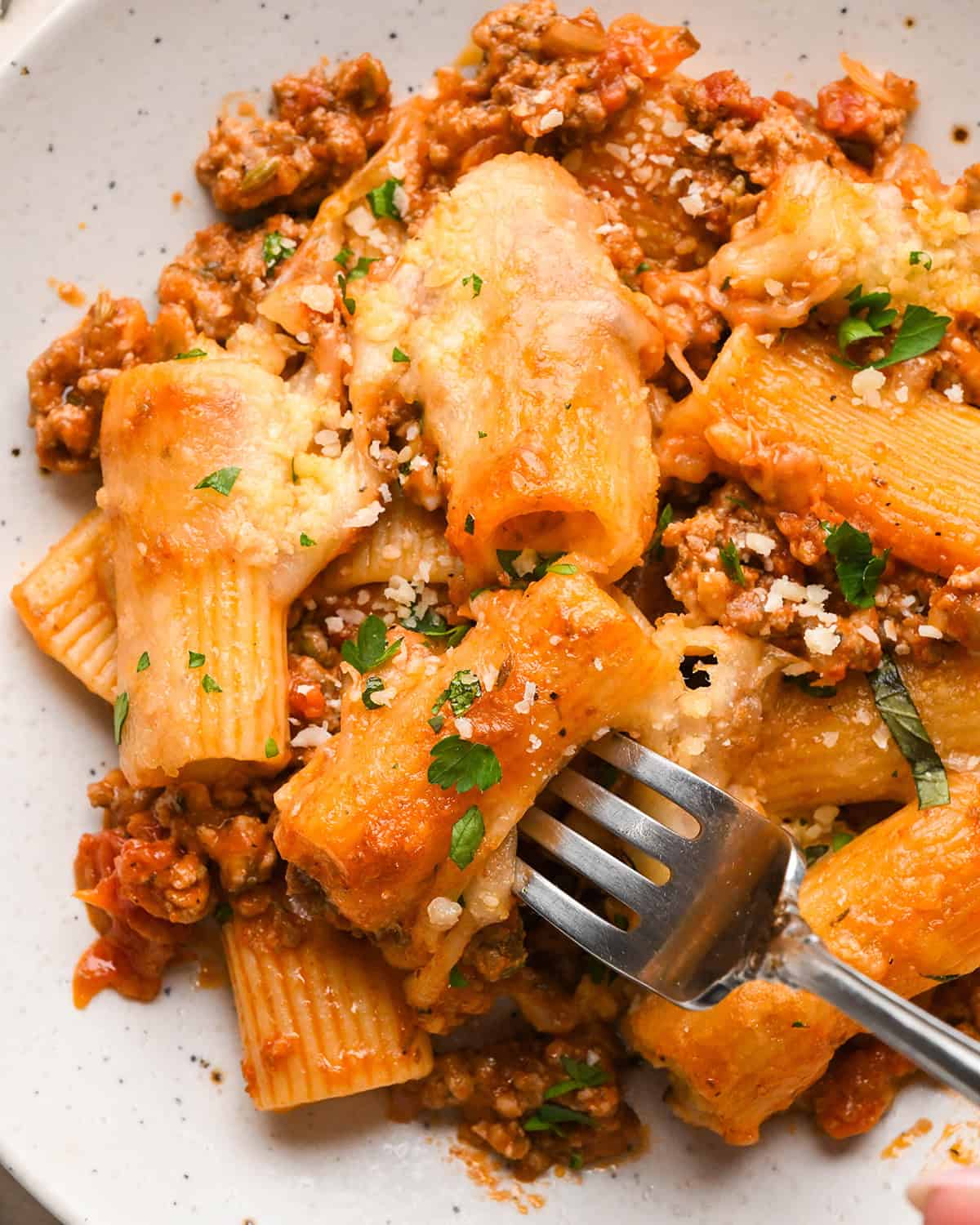 Close-up photo of baked ziti topped with fresh herbs, served on a white speckled plate with a fork.