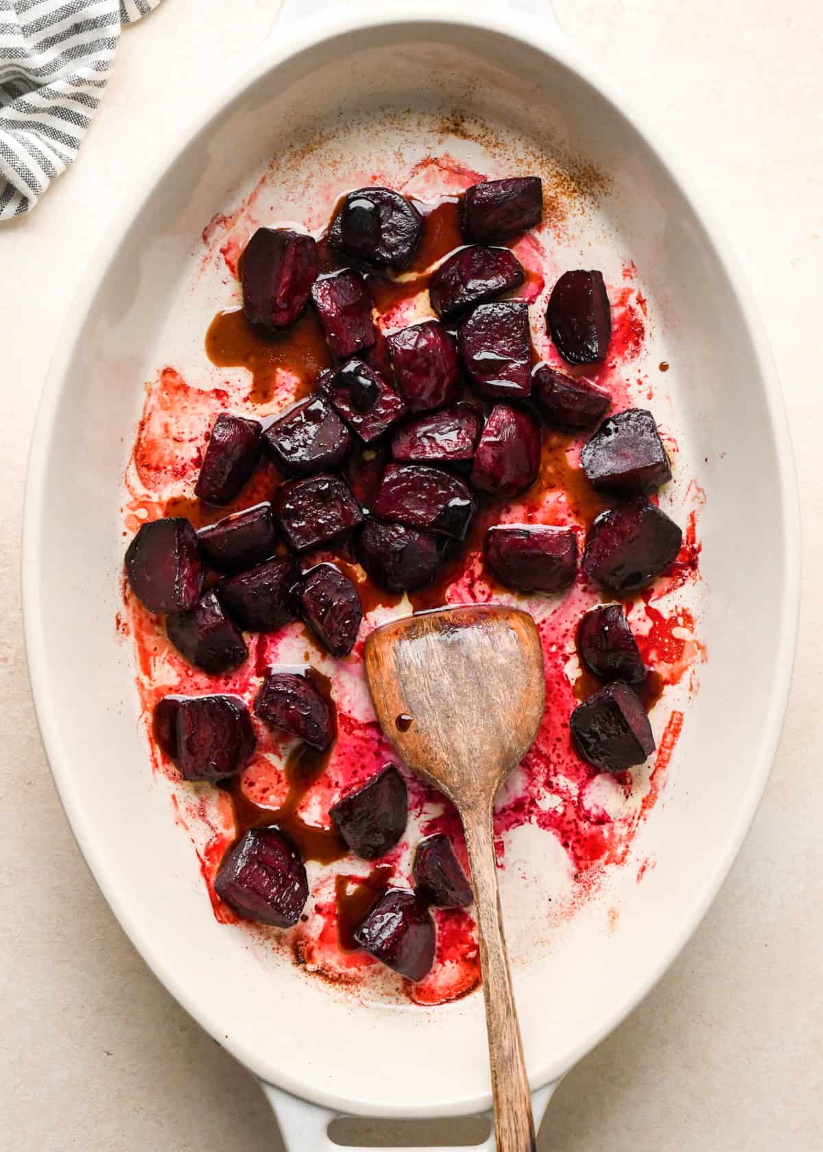 balsamic honey mixture added to roasted beets in a white oval ceramic baking dish with a wooden spatula