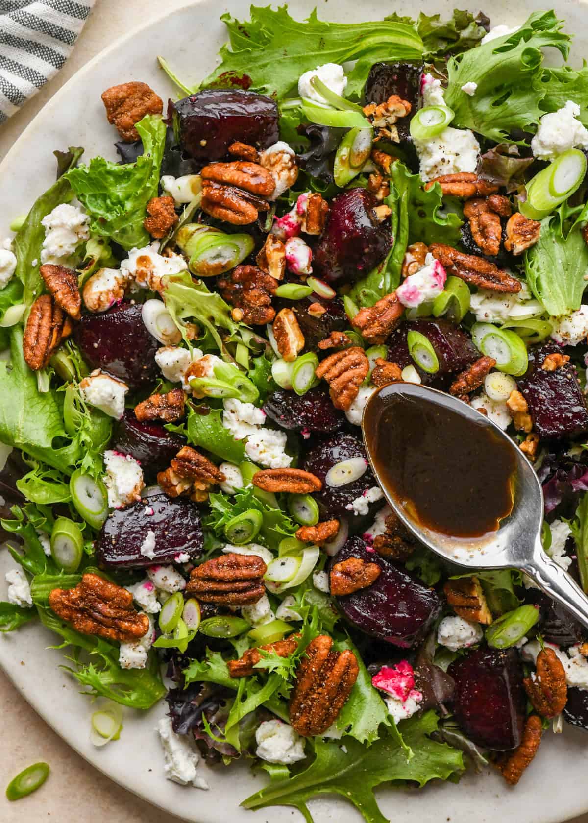 dressing being poured over beet salad on a white plate, made with mixed greens, roasted beets, goat cheese, sliced green onions, and candied pecans.