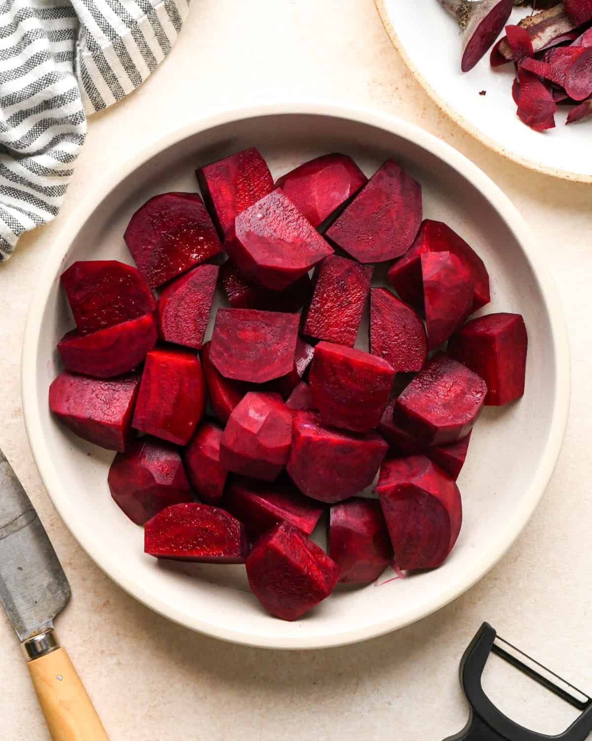 peeled and cut beets in a white bowl on a tan surface with a knife and a peeler nearby.