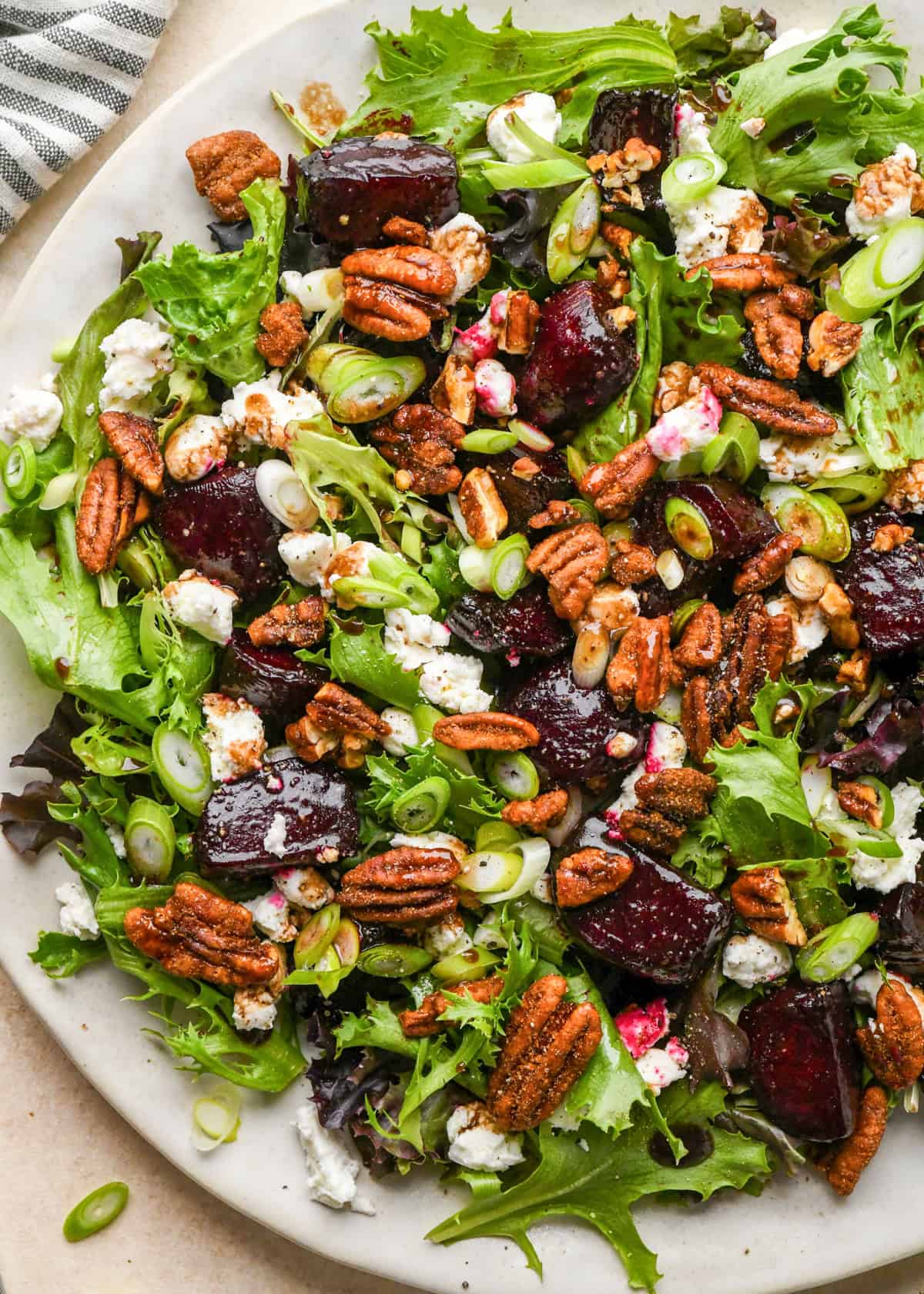 close-up of a beet salad on a white plate, made with mixed greens, roasted beets, goat cheese, sliced green onions, and candied pecans.