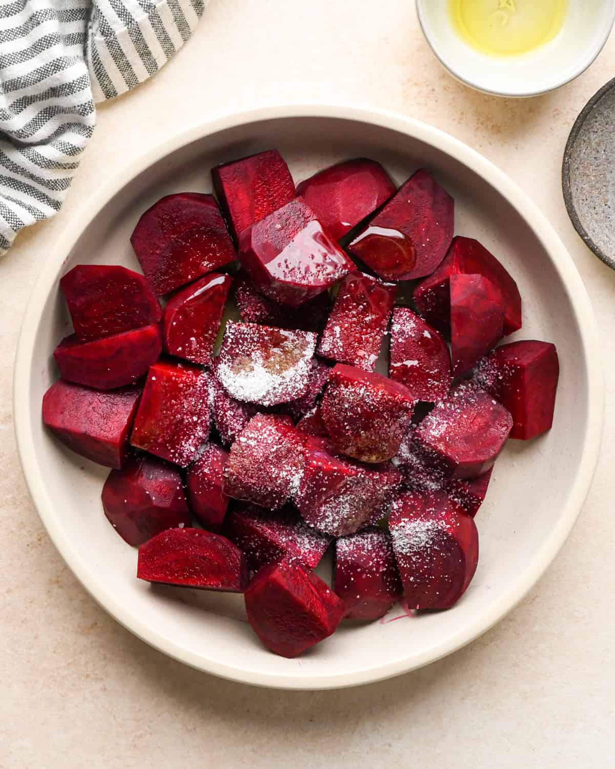 olive oil, salt and pepper added to peeled and cut beets in a white bowl on a tan surface before mixing