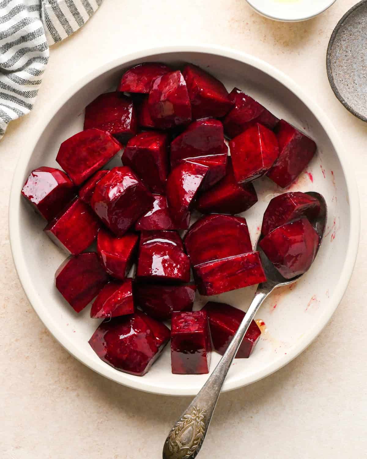 olive oil, salt and pepper added to peeled and cut beets in a white bowl on a tan surface after mixing with a silver spoon