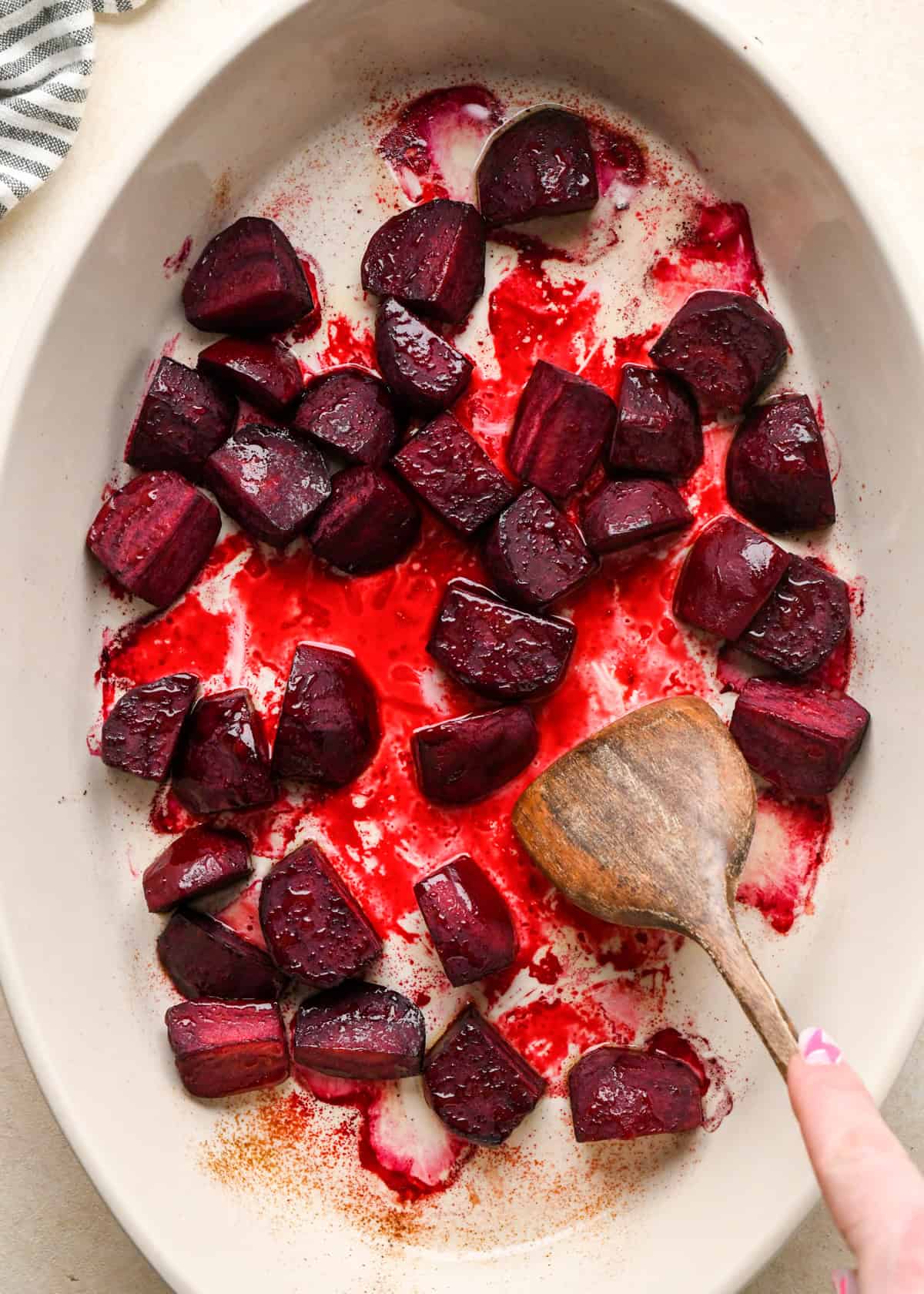 a wooden spatula stirring beets in a white, oval ceramic baking dish to be used in a beet salad