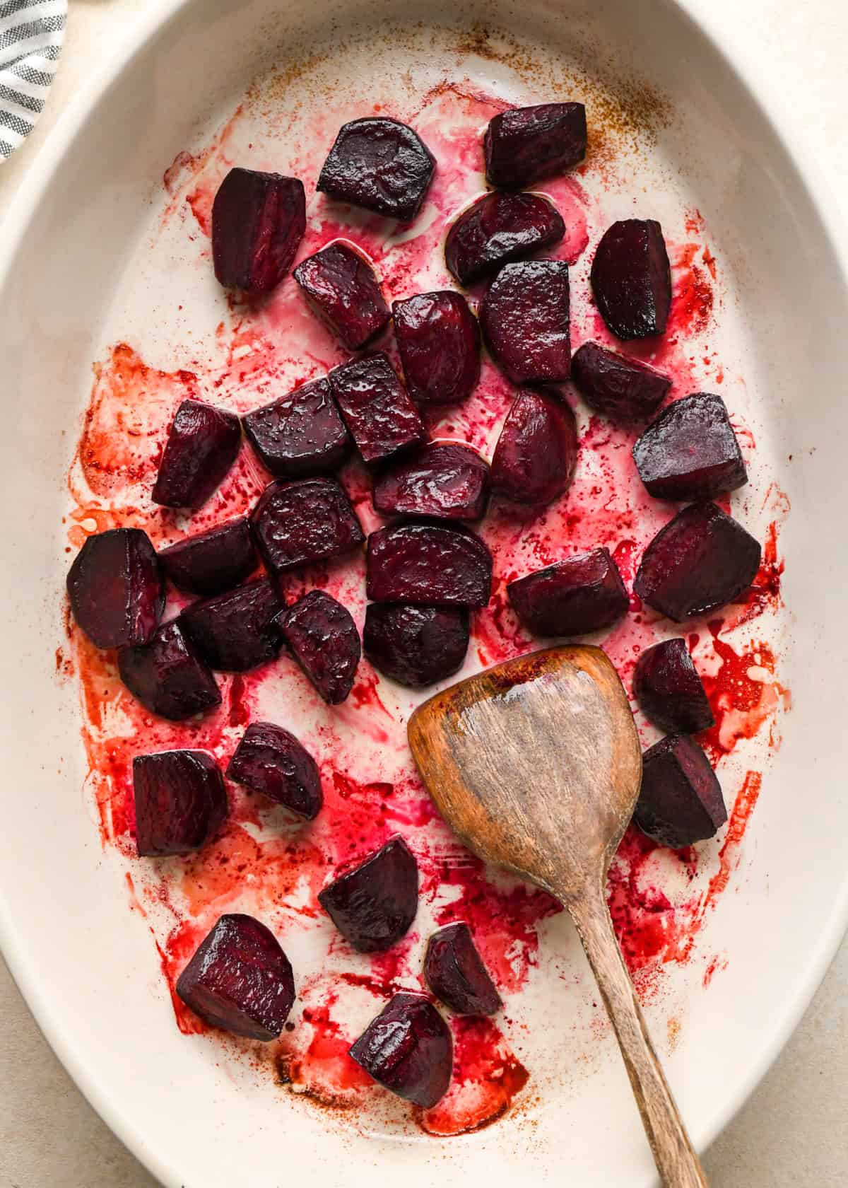a wooden spatula stirring beets in a white, oval ceramic baking dish to be used in a beet salad