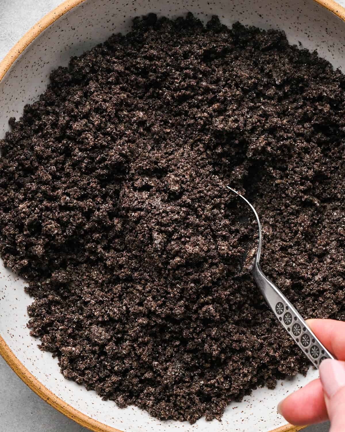 a hand holding a silver spoon stirring the oreo crumbs in a ceramic bowl making the crust for chocolate peanut butter pie