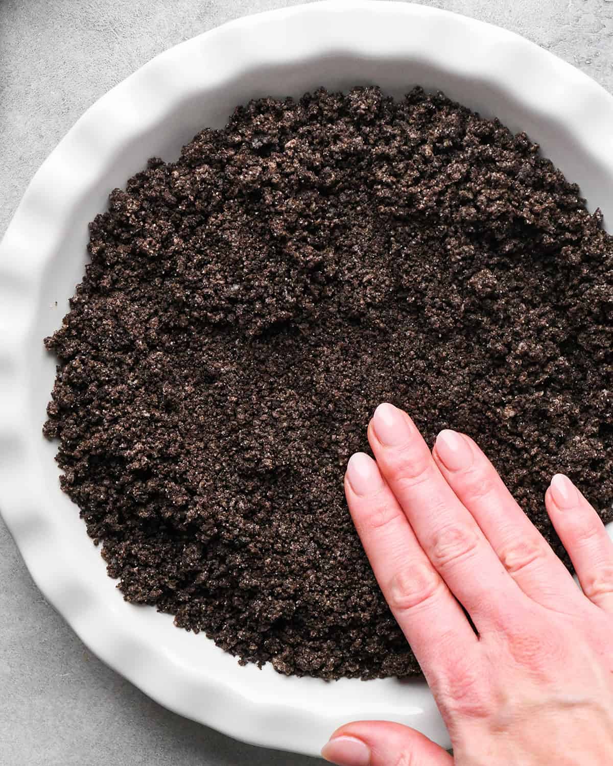 a hand pressing the oreo crust mixture into a white pie dish making chocolate peanut butter pie