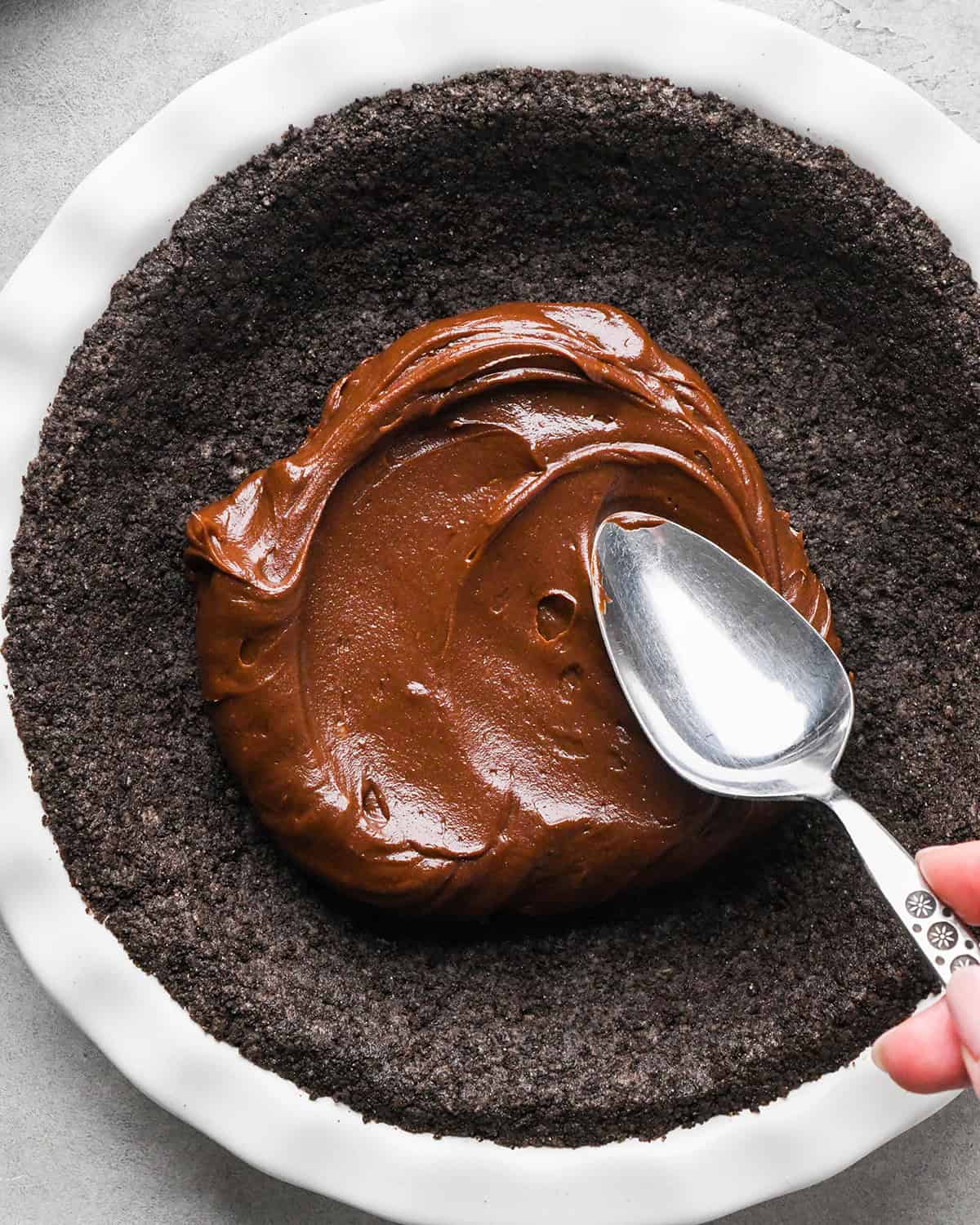 chocolate ganache being spread over the baked oreo crust with a silver spoon making chocolate peanut butter pie
