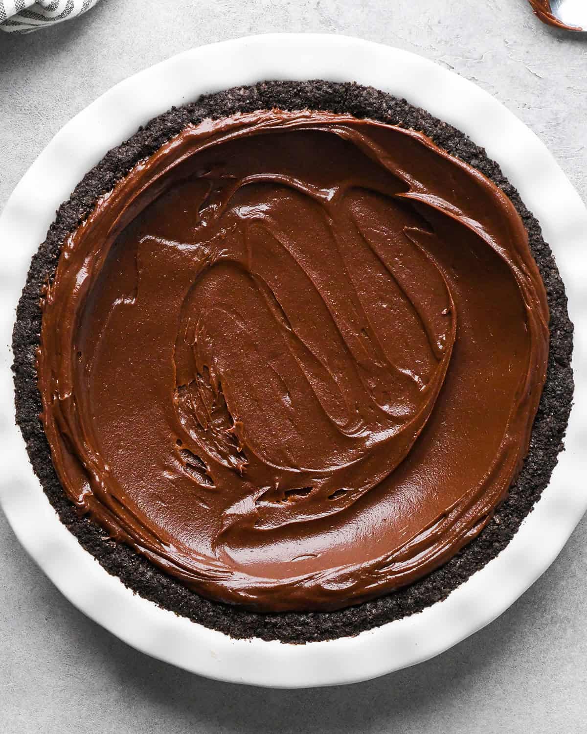 chocolate ganache evenly spread over the baked oreo crust with a silver spoon making chocolate peanut butter pie