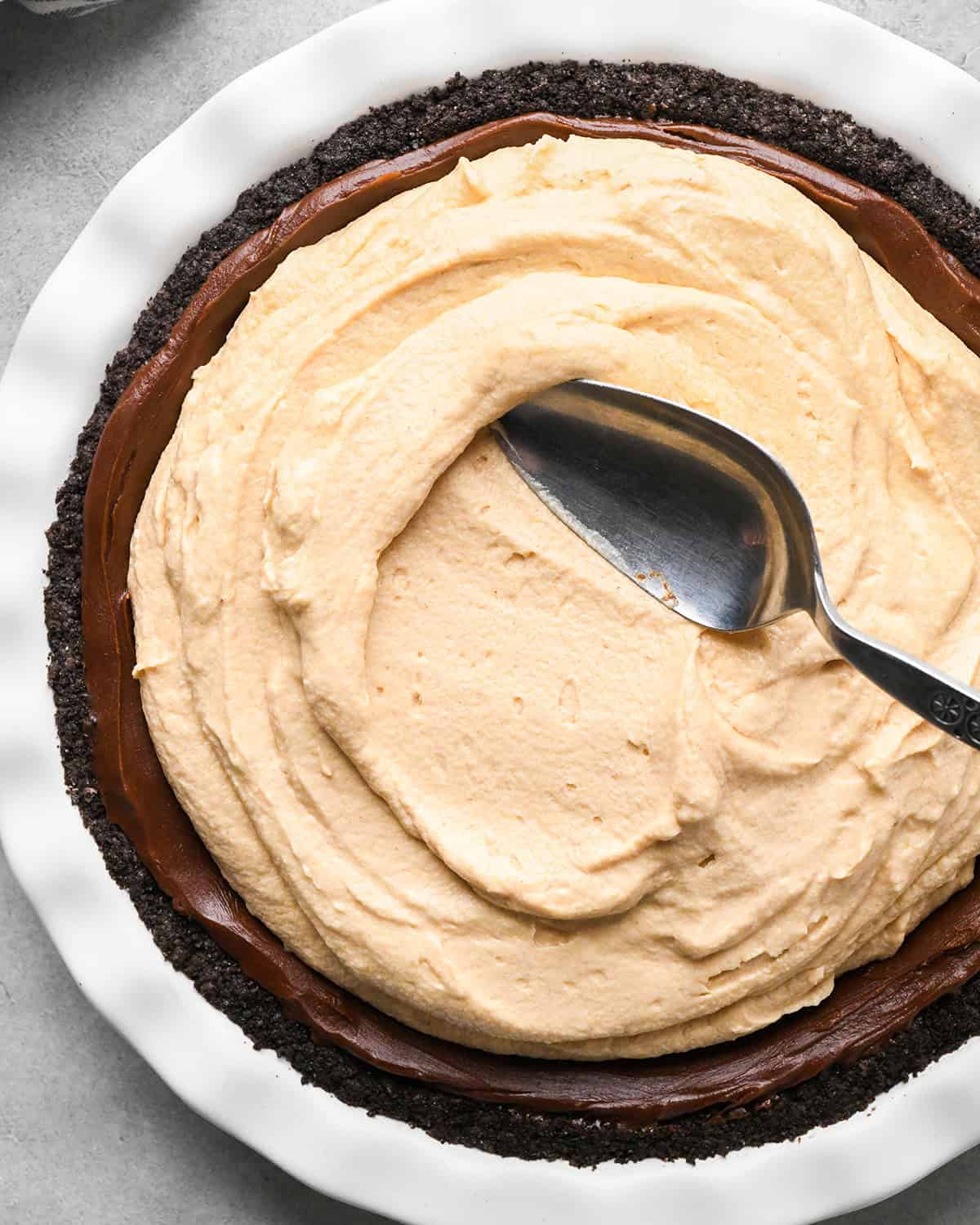 peanut butter filling being spread with a silver spoon over the chocolate ganache layer of a chocolate peanut butter pie in a white pie dish