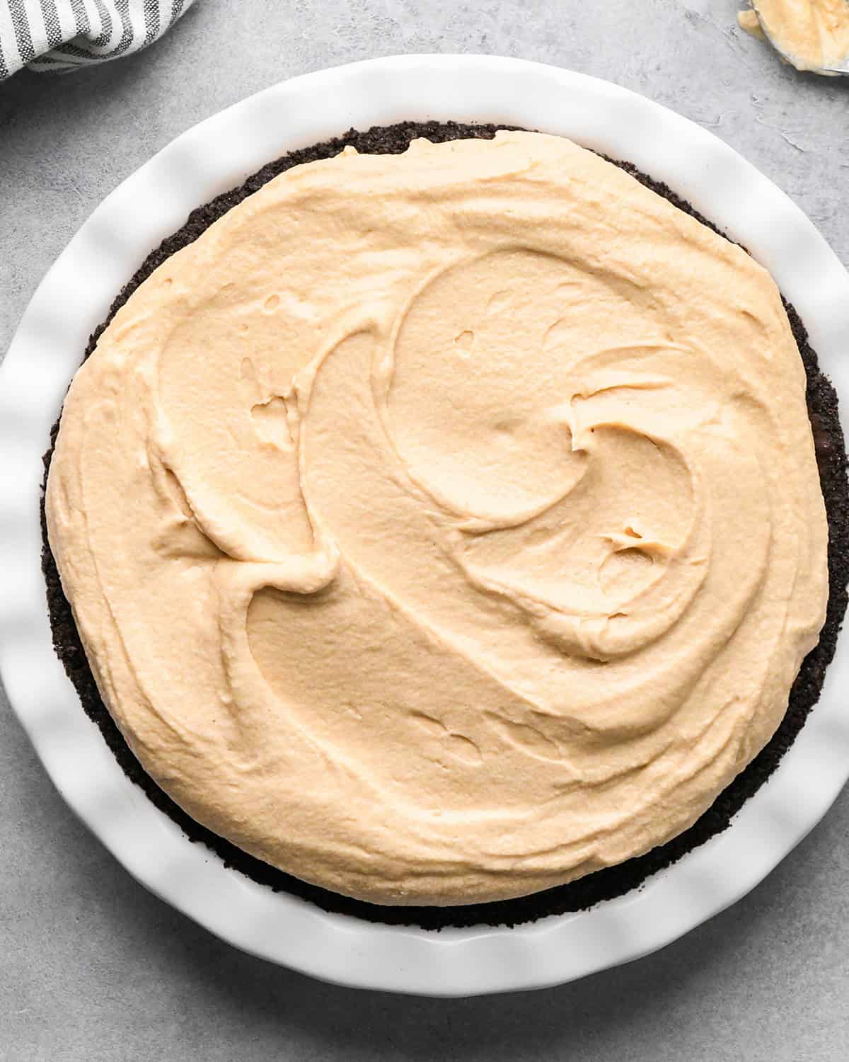 peanut butter being spread over the chocolate ganache layer of a chocolate peanut butter pie in a white pie dish on a gray surface