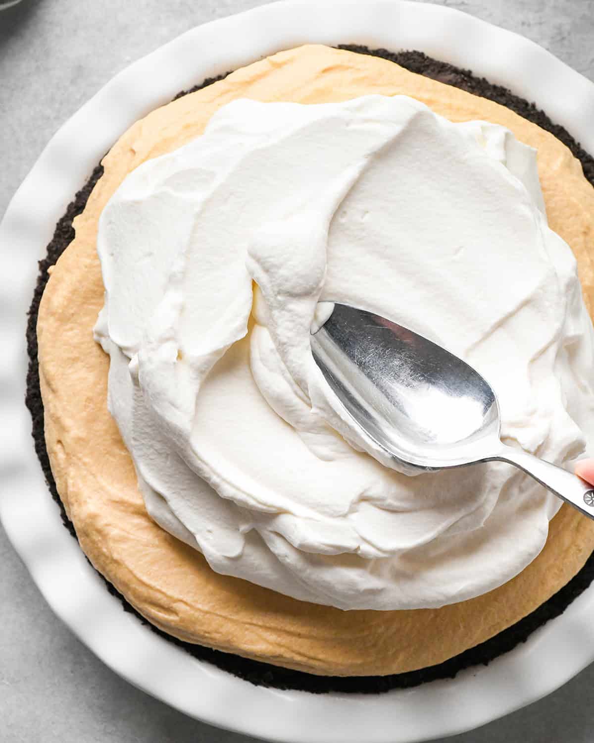 whipped cream being spread with a silver spoon over the peanut butter layer of this chocolate peanut butter pie recipe