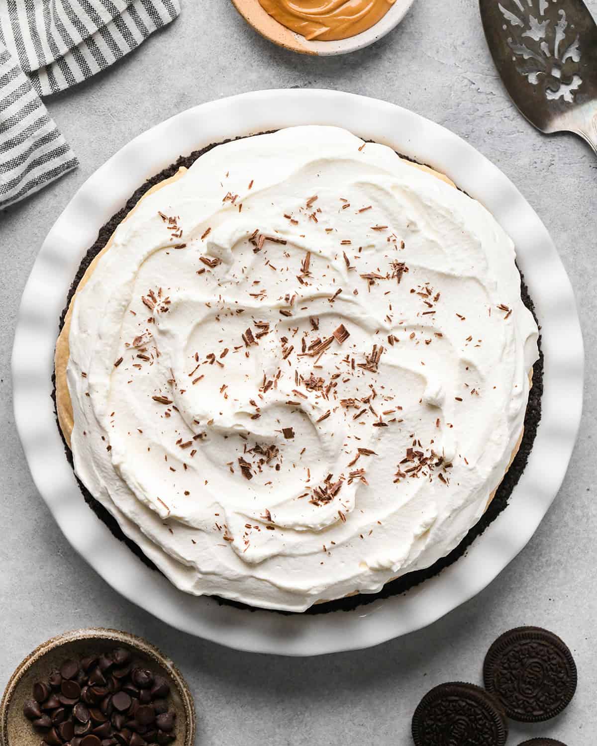 chocolate peanut butter pie with chocolate shavings on top on a gray surface in a white pie dish surrounded by chocolate chips, oreos and a pie server