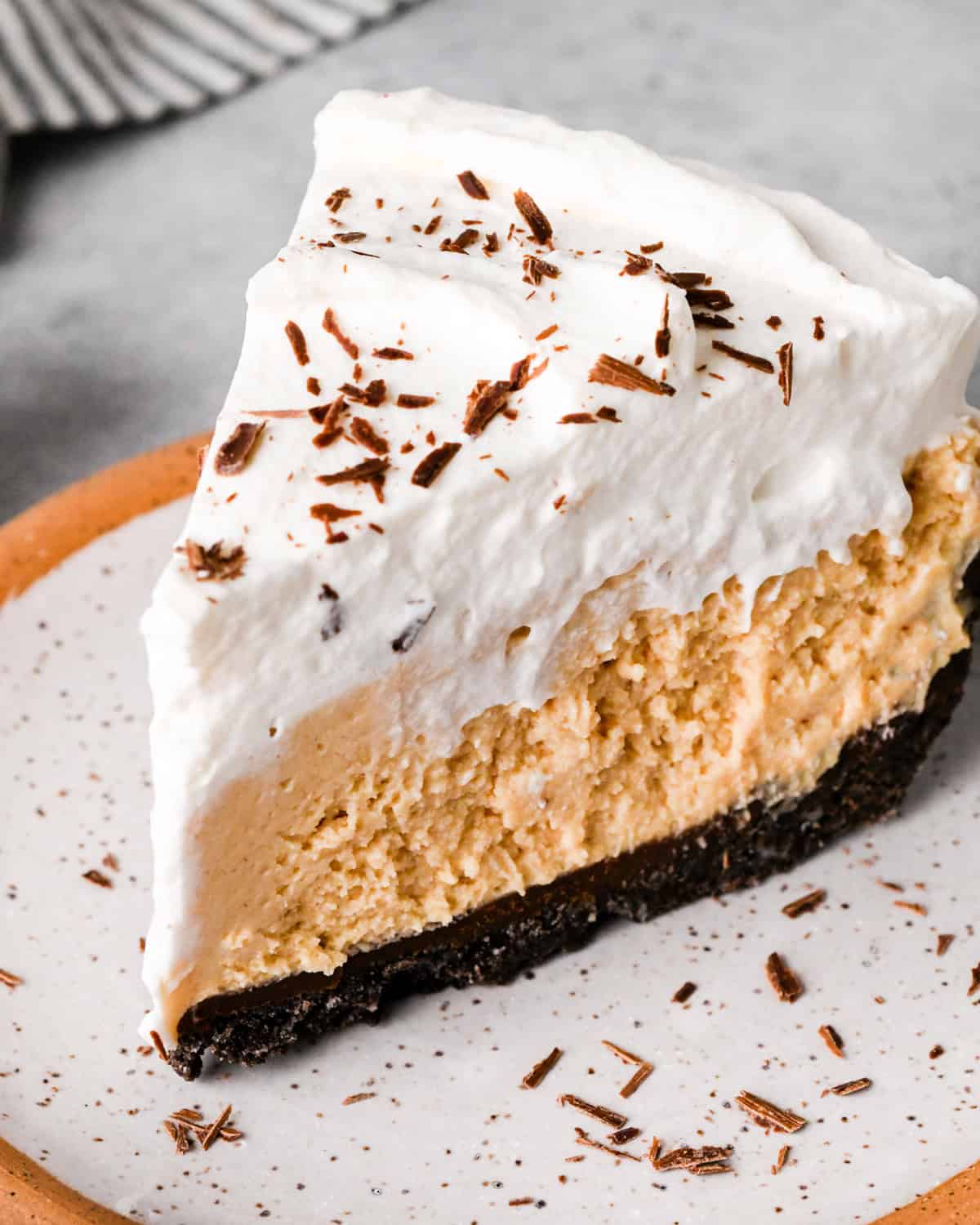 a slice of chocolate peanut butter pie on a speckled ceramic plate. The pie has 4 layers from bottom to top they are an oreo cookie crust, chocolate ganache, peanut butter pie filling and whipped cream with chocolate shavings.