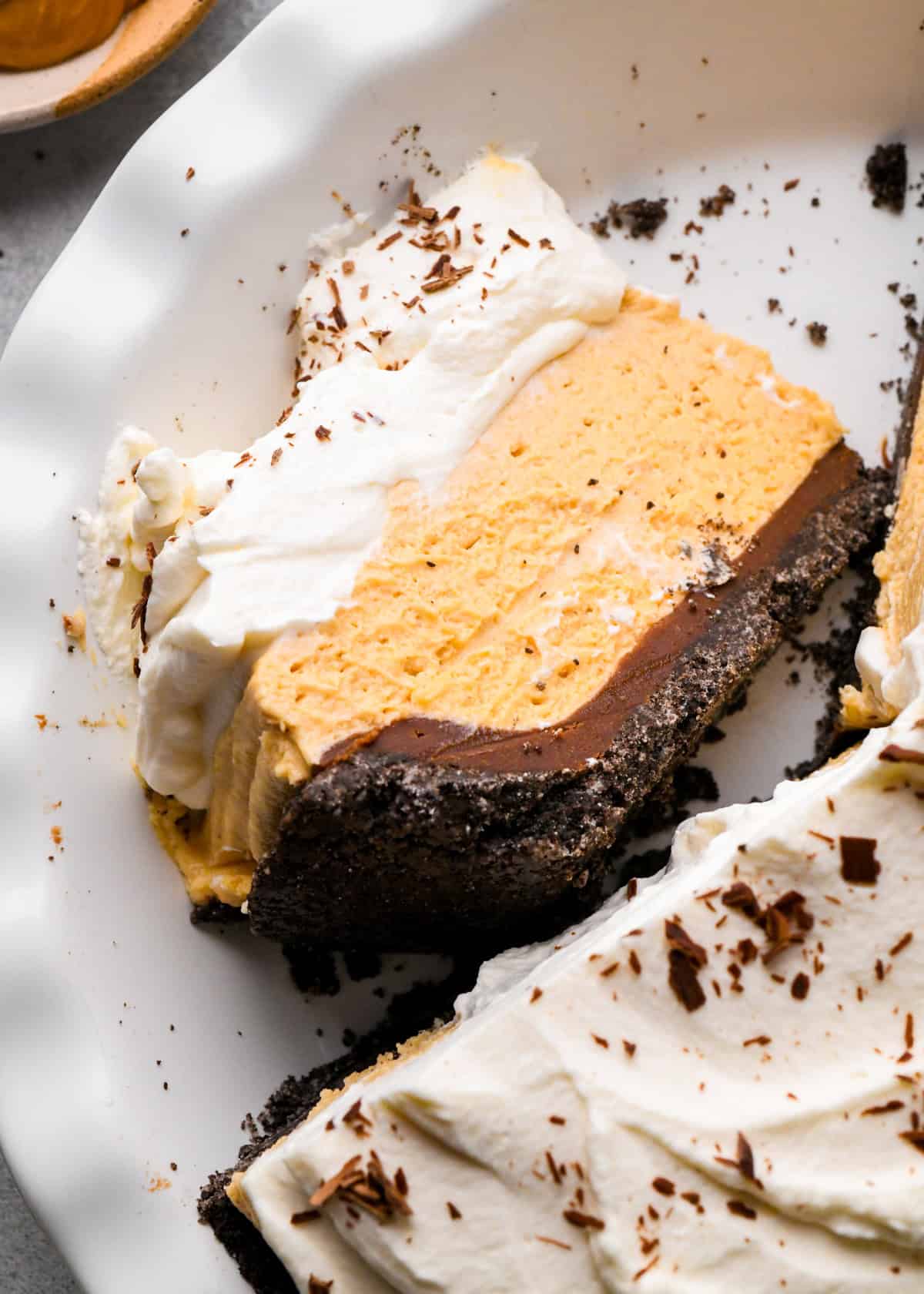 a slice of chocolate peanut butter pie in a pie dish on it's side. The pie has 4 layers from bottom to top they are an oreo cookie crust, chocolate ganache, peanut butter pie filling and whipped cream with chocolate shavings. The rest of the pie is also in the dish.