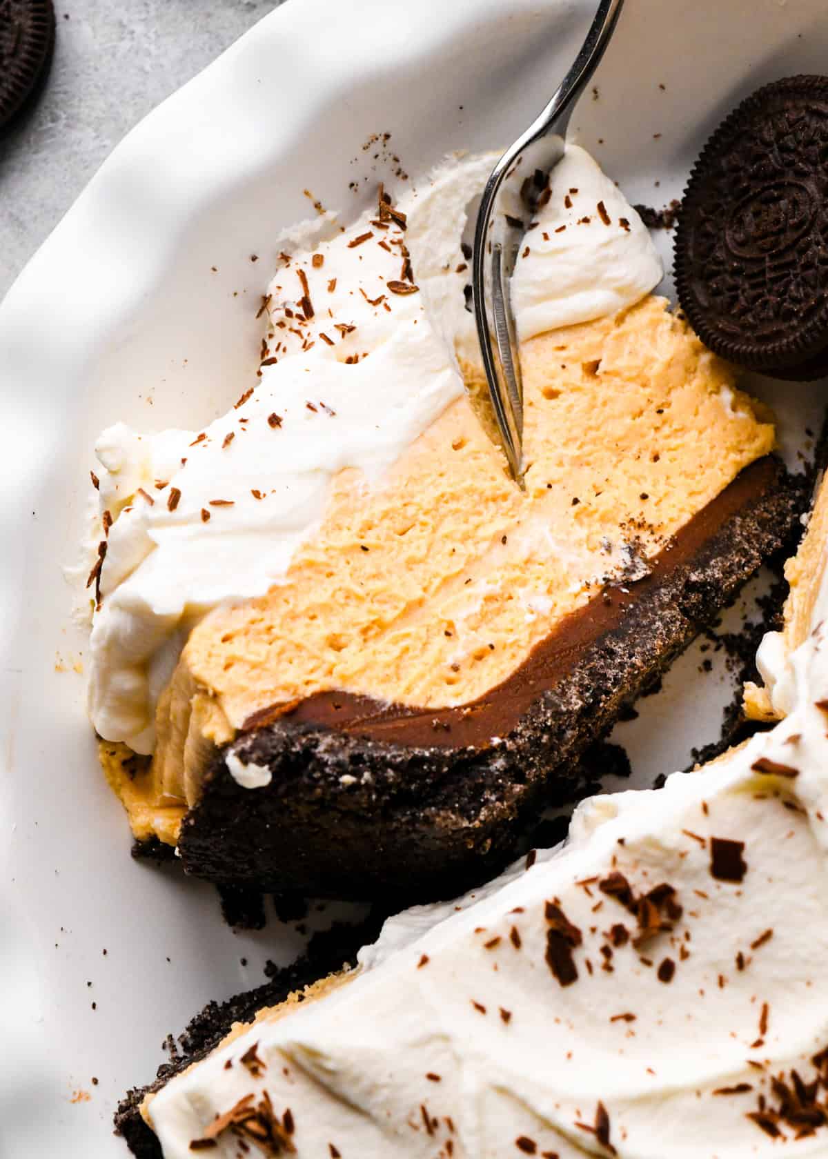 a slice of chocolate peanut butter pie in a pie dish on it's side with a fork taking a bite. The pie has 4 layers from bottom to top they are an oreo cookie crust, chocolate ganache, peanut butter pie filling and whipped cream with chocolate shavings. The rest of the pie is also in the dish with an oreo cookie.