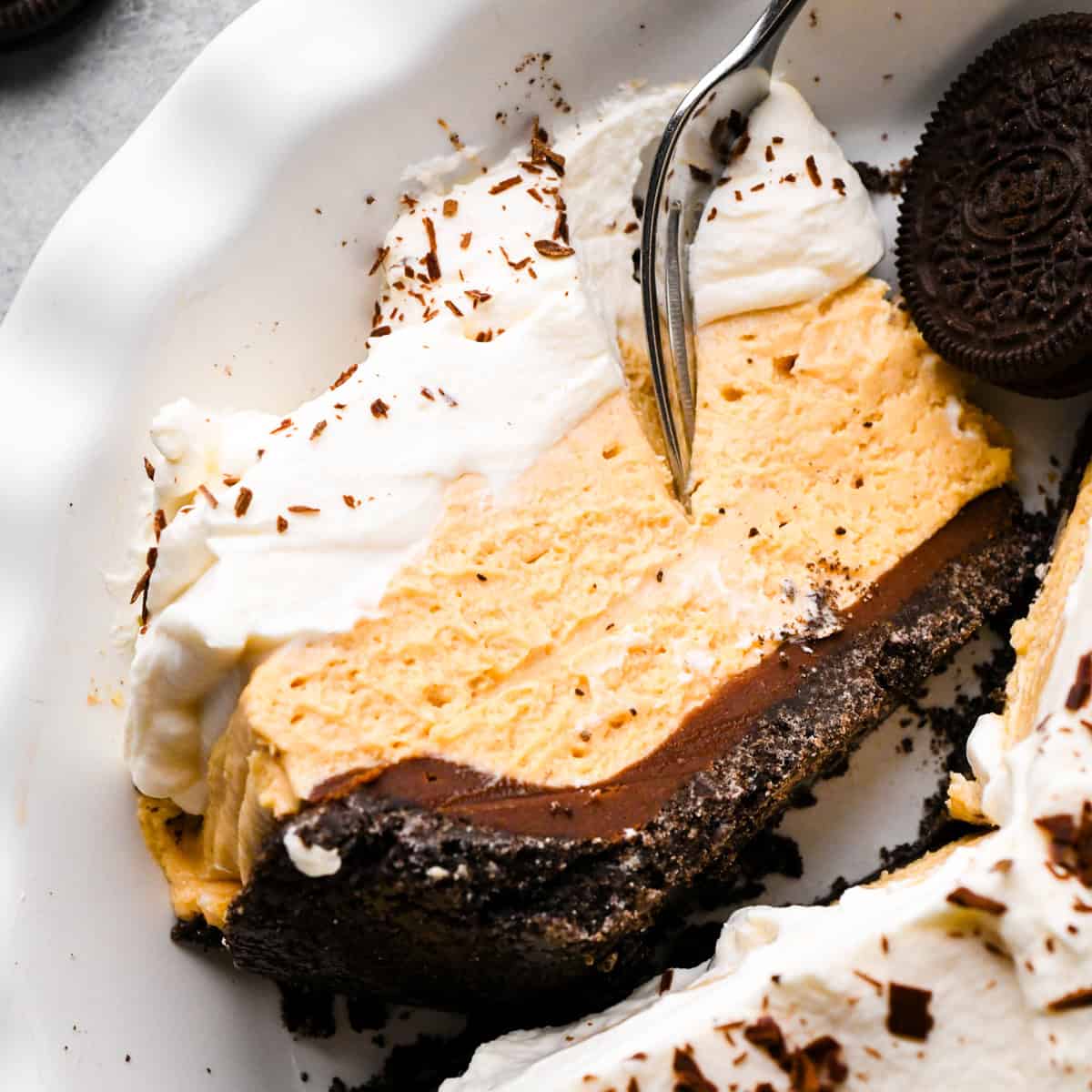 a slice of chocolate peanut butter pie in a pie dish on it's side with a fork taking a bite. The pie has 4 layers from bottom to top they are an oreo cookie crust, chocolate ganache, peanut butter pie filling and whipped cream with chocolate shavings. The rest of the pie is also in the dish with an oreo cookie.
