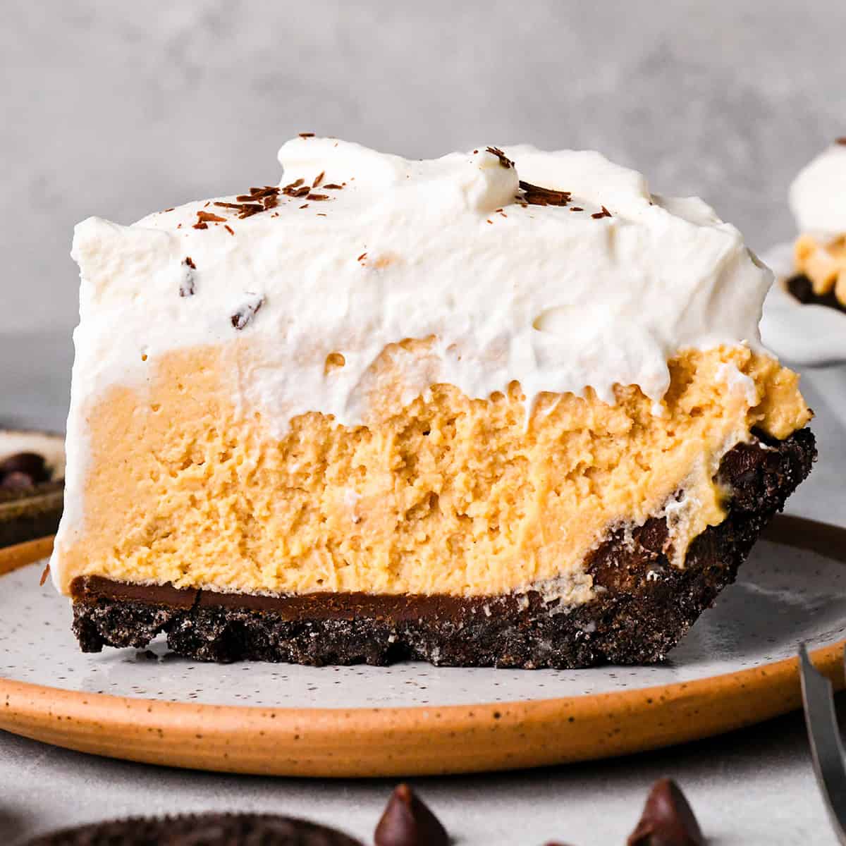 a slice of chocolate peanut butter pie on a speckled ceramic plate. The pie has 4 layers from bottom to top they are an oreo cookie crust, chocolate ganache, peanut butter pie filling and whipped cream with chocolate shavings. There are chocolate chips and cookies around the plate.