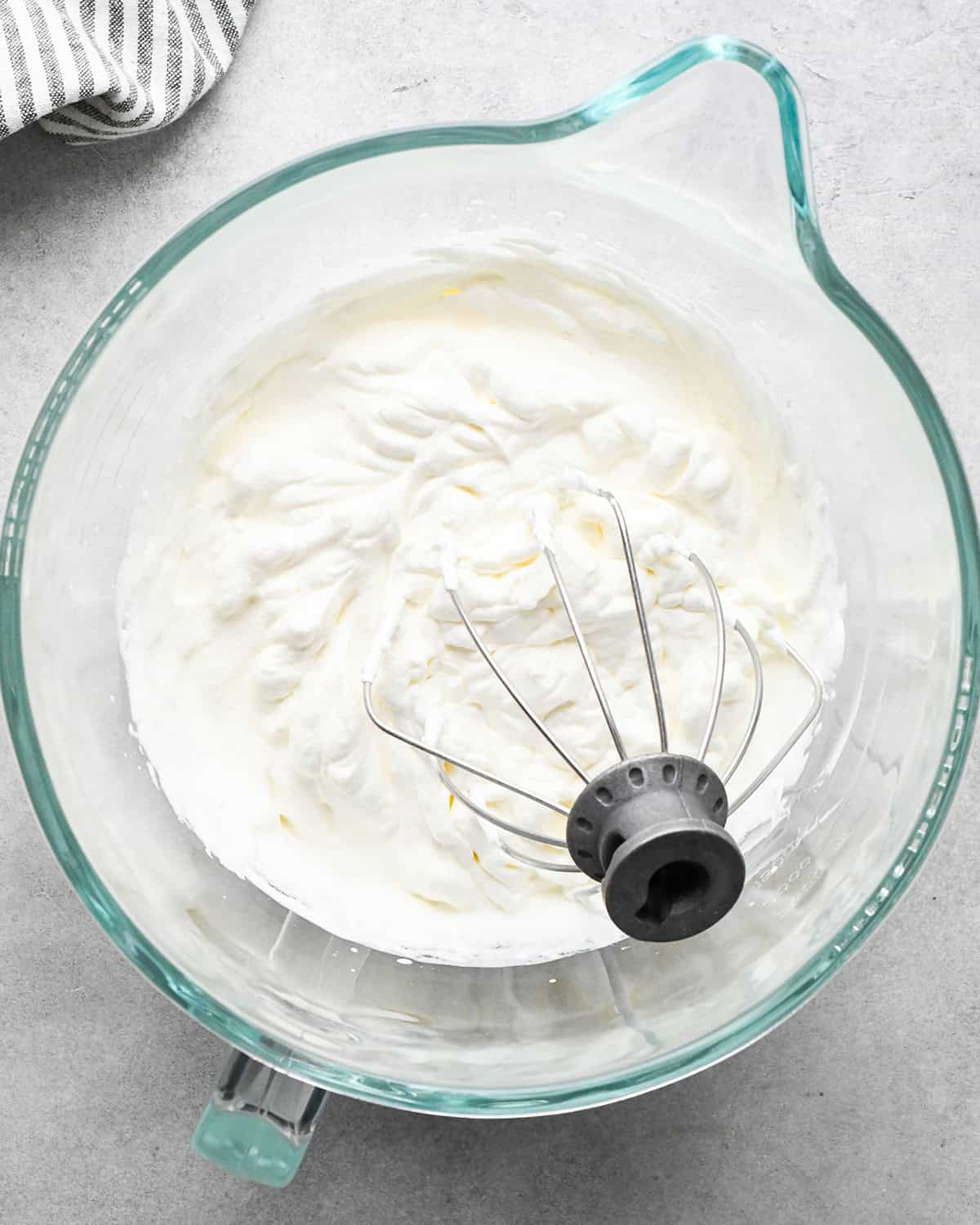 whipped heavy cream in a glass mixing bowl with the wire whisk attachment in the bowl on a gray surface