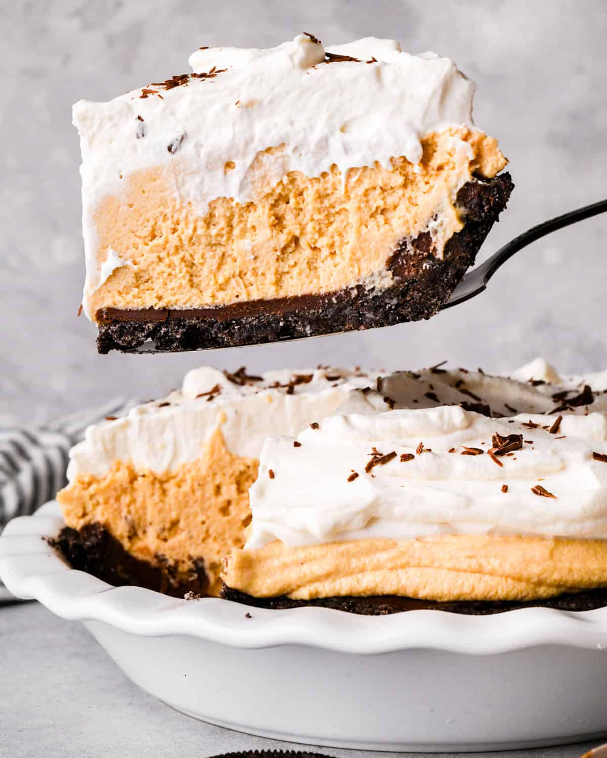 a slice of chocolate peanut butter pie on a pie server being held over the pie in a pie dish. The pie has 4 layers from bottom to top they are an oreo cookie crust, chocolate ganache, peanut butter pie filling and whipped cream with chocolate shavings.