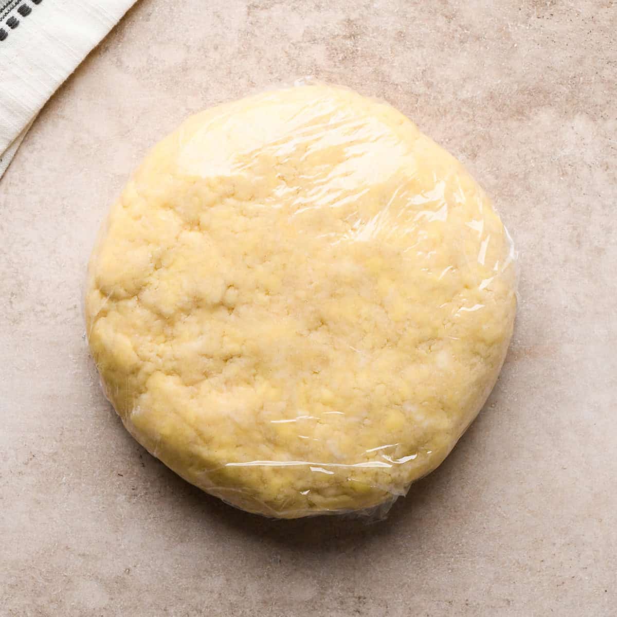 a disc of pie dough wrapped in plastic wrap on a tan surface