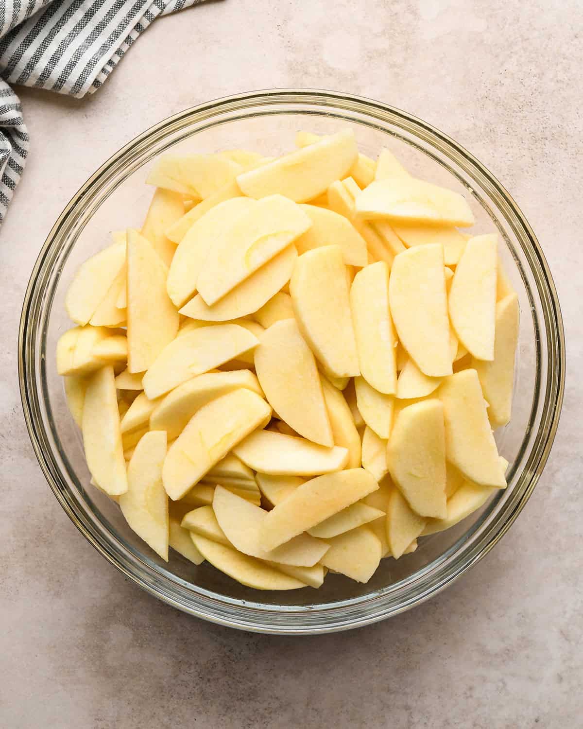 peeled and sliced apples in a glass bowl before cooking on a tan surface with a striped towel in the top left corner