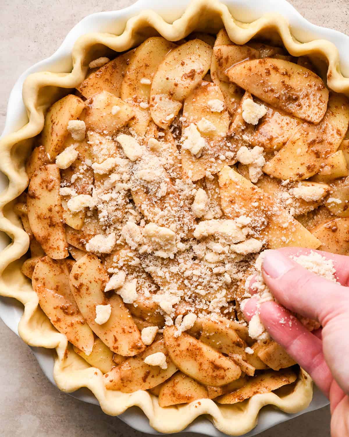 a hand sprinkling the crumb topping over dutch apple pie filling in a pie crust with a fluted edge in a white ceramic pie dish