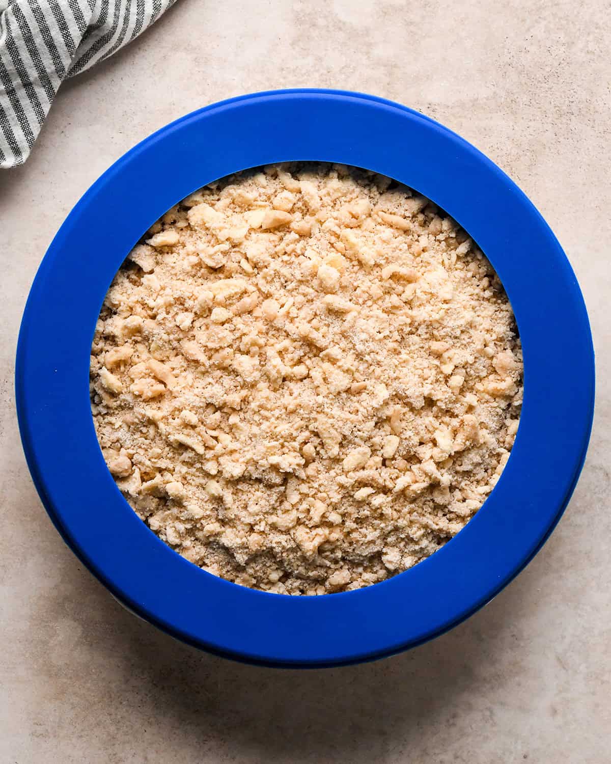 dutch apple pie in a pie dish with a blue silicone pie crust shield around the edge on a tan surface with a striped towel in the upper left corner before baking