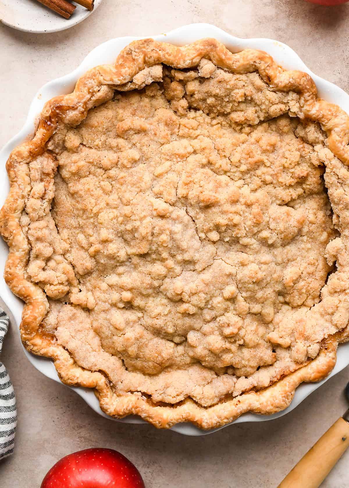 dutch apple pie in a white ceramic pie dish on a tan surface with a striped towel and cinnamon sticks in the background