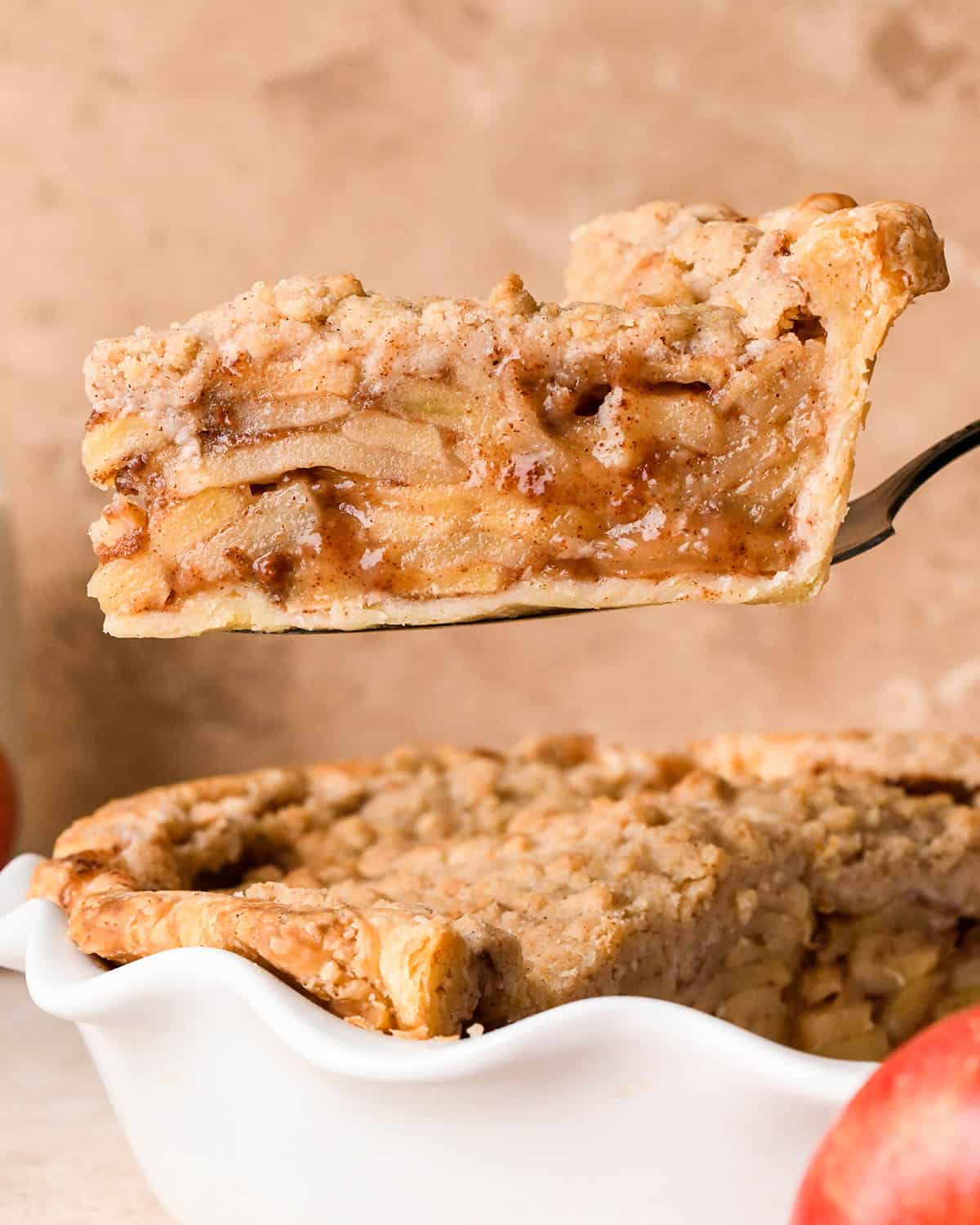 a piece of dutch apple pie being lifted out of a pie dish with a pie server - showing the delicious layers of apple pie filling between the bottom crust and crumb topping