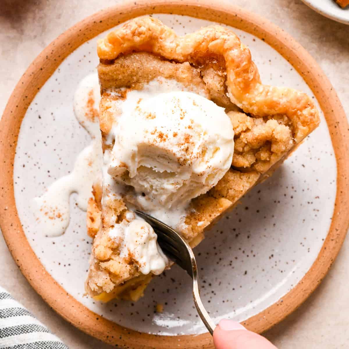 a slice of dutch apple pie on a speckled ceramic plate topped with vanilla ice cream and a sprinkle of cinnamon. There's a fork taking a bite of the pie