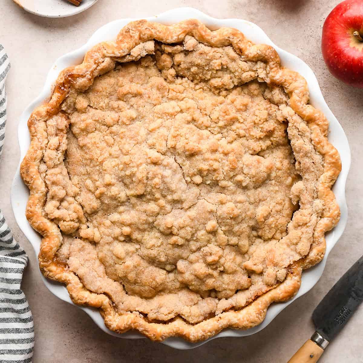 dutch apple pie in a white ceramic pie dish on a tan surface surrounded by an apple, cinnamon sticks and a striped towel.