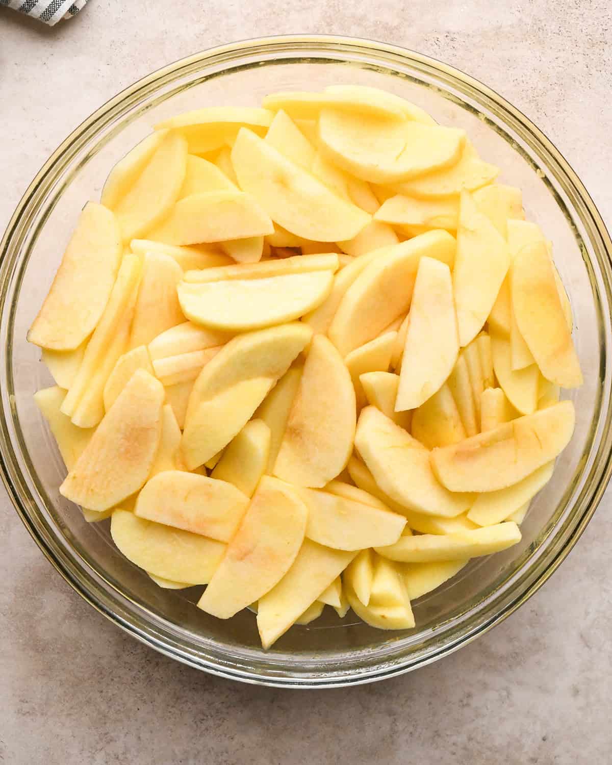 peeled and sliced apples in a glass bowl after cooking on a tan surface with a striped towel in the top left corner