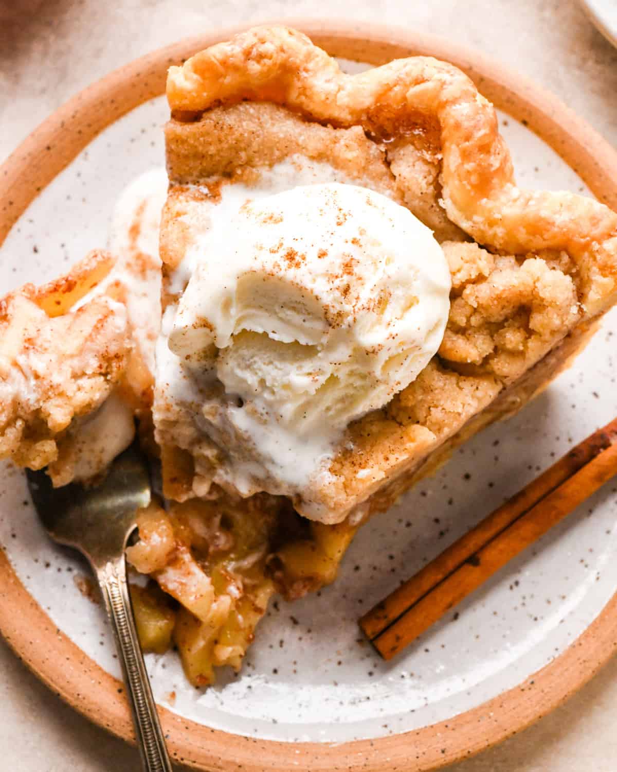 a slice of dutch apple pie on a ceramic plate topped with ice cream and cinnamon. There's a fork on the plate with a bite on it and a cinnamon stick.