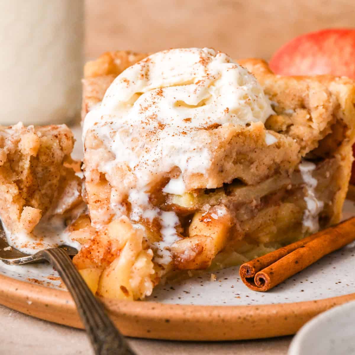 front view of a slice of dutch apple pie on a speckled ceramic plate topped with vanilla ice cream and a sprinkle of cinnamon. There's a fork taking a bite of the pie with an apple and cinnamon stick nearby