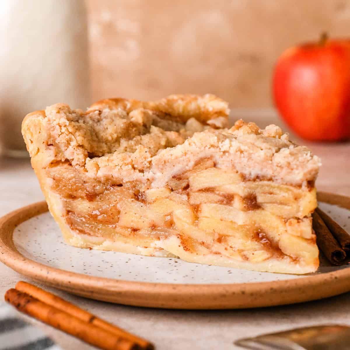 A slice of Dutch apple pie on a speckled ceramic plate, with layers of apple filling and a crumb topping. A fresh red apple and cinnamon sticks are blurred in the background.