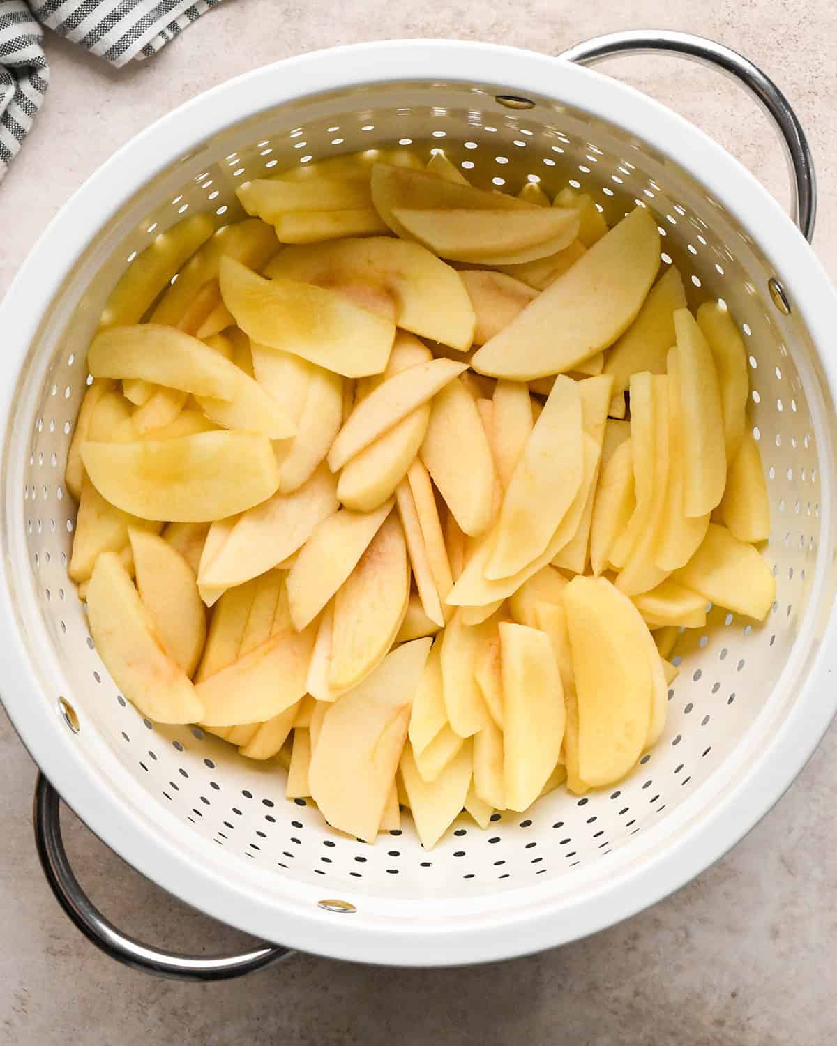 peeled and sliced apples in a colander after cooking on a tan surface with a striped towel in the top left corner