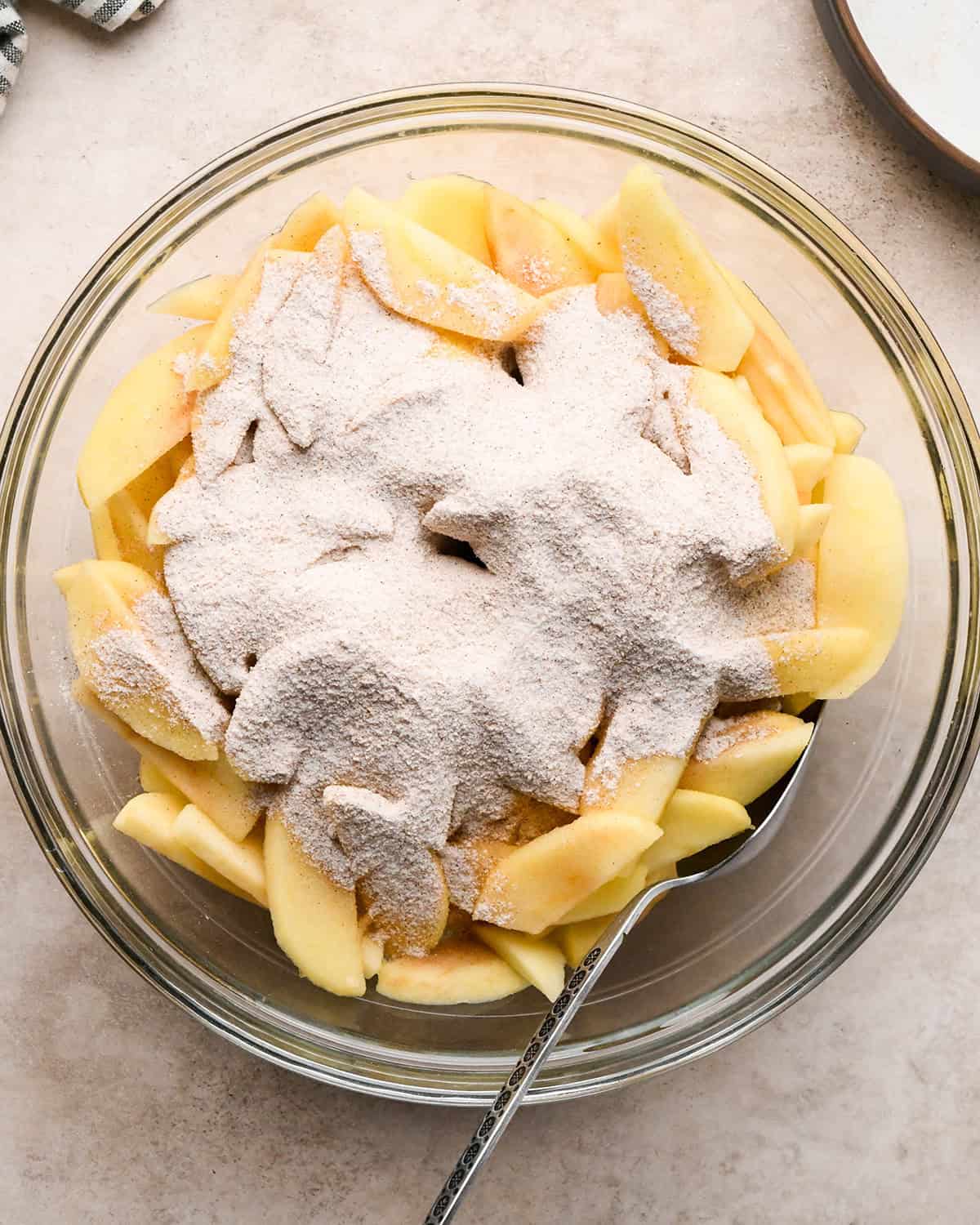 dry ingredient mixture added to peeled, sliced and cooked apples in a glass bowl before mixing with a silver spoon in it on the side on a tan surface making dutch apple pie