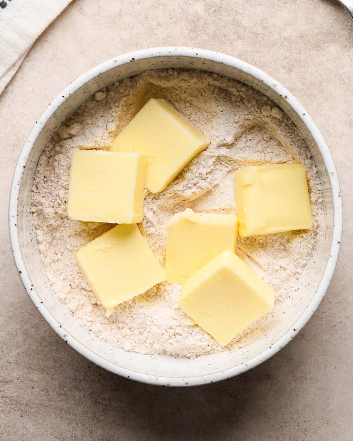 cubes of butter added to the flour mixture in a white speckled bowl before mixing on a tan surface making the crumb topping for dutch apple pie