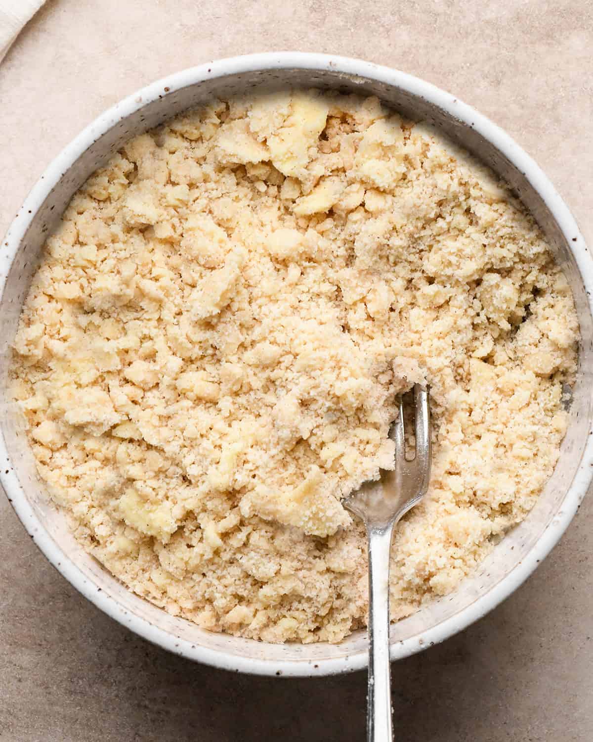 dutch apple pie crumb topping in a white speckled ceramic bowl with a silver fork on a tan surface