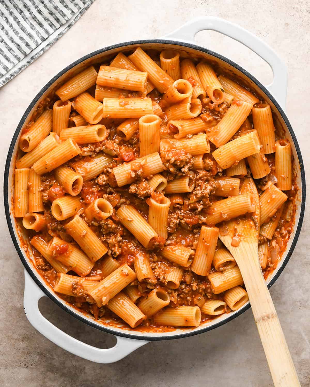 Making baked ziti - cooked ziti pasta is added to saucepan with the prepared sauce, and ingredients are being stirred together with a wooden spoon.